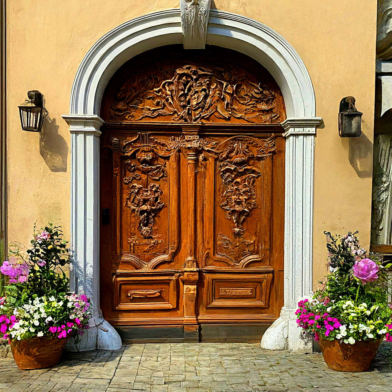 Ornate carved wooden door with white arch frame and flower pots on yellow wall