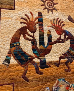 Art quilt with two Kokopelli figures, saguaro cactus, and sun motif in earth tones with turquoise geometric patterns