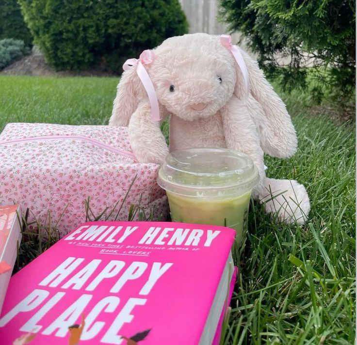 Plush bunny on grass next to a pink book and a cup of green drink outdoors