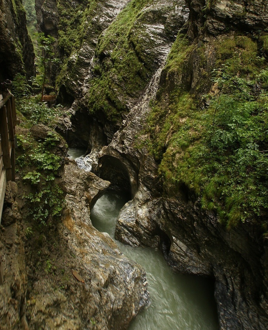 Alpine waterfall cascading down rocky cliffs into a stream with green mountains in the background.
