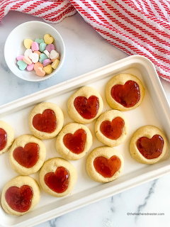 Heart-shaped jam cookies on white tray with candies in bowl on red striped cloth