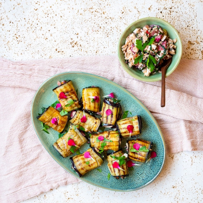 Plate of aubergine rolls with herbs and pomegranate seeds beside a bowl of orzo salad