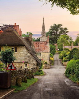 Quaint English village street with thatched cottage, church spire, and green hedges at sunset