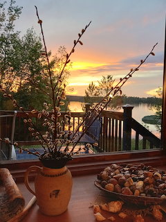 Pussy willow branches in a ceramic pitcher on a deck railing overlooking a lake at sunset