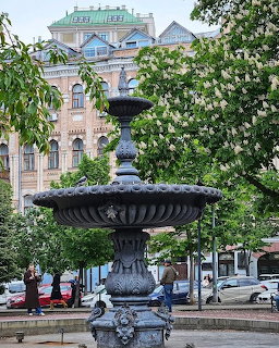 Ornate fountain in a city square with blooming trees and a historic building behind it