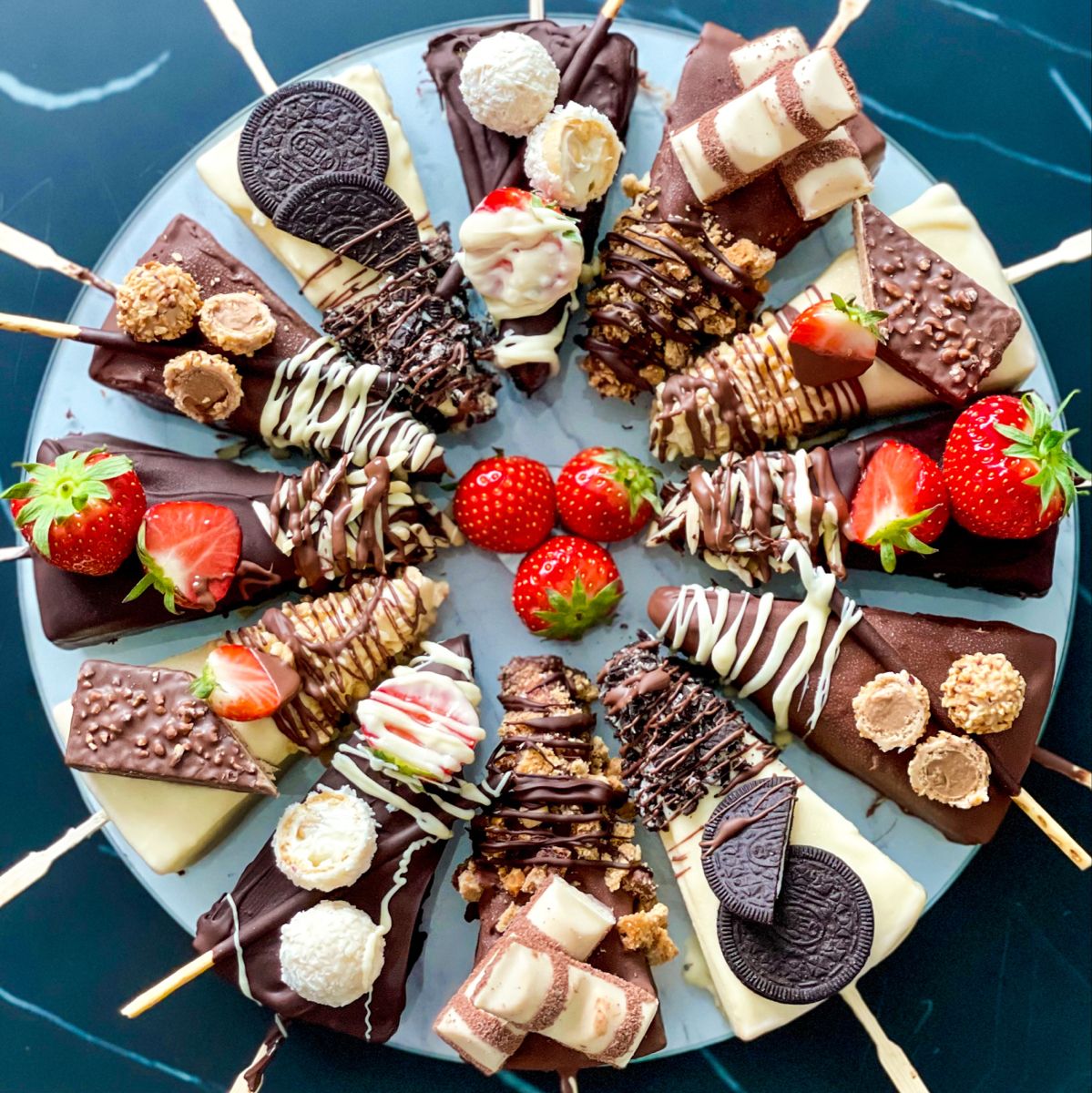 Chocolate-covered cheesecake sticks on a platter with strawberries, cookies, and assorted sweet toppings