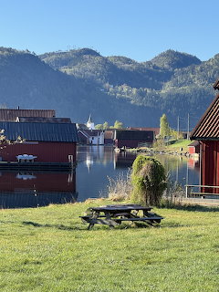 Red wooden buildings along a calm fjord harbor with mountains, green lawn and picnic table in foreground