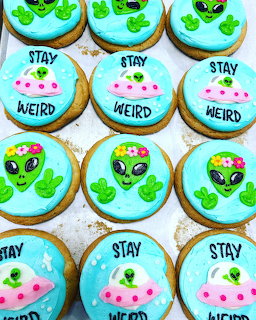 Sugar cookies decorated with green aliens and pink UFOs with 'STAY WEIRD' text on blue icing