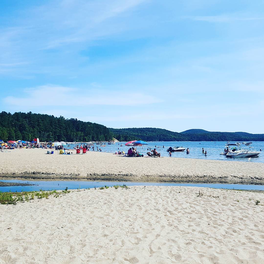 Sandy beach with people, calm water, forested hills and blue sky at Sandy Hook, Iles-de-la-Madeleine