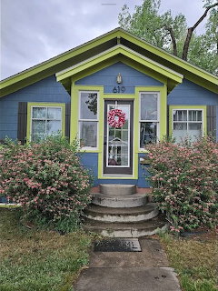 Small blue cottage with yellow trim, front steps, shrubs, and a wreath on the glass door