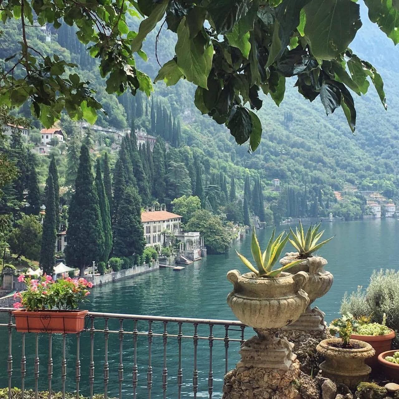 Ornate stone planter on terrace overlooking blue lake with mountains and villa, framed by foliage