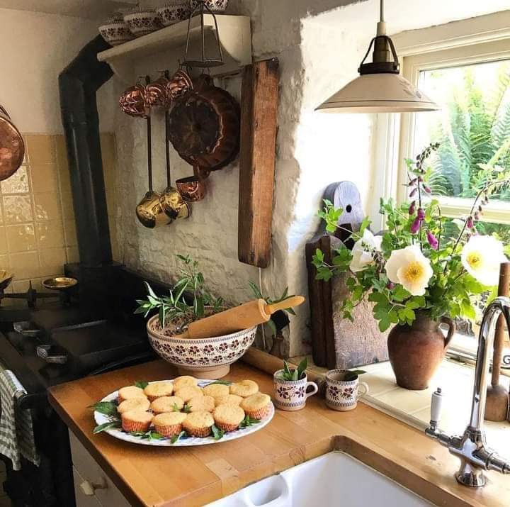 Rustic kitchen interior with black stove, wooden counter, fresh flowers in vases, and plate of scones by sunny window