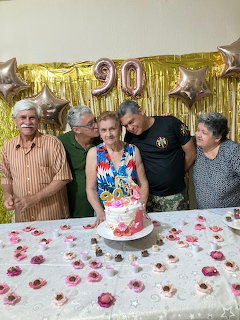 Five adults posing behind a birthday cake with gold balloons and a 90 decoration indoors.