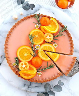 Orange citrus tart garnished with orange slices, kumquats, rosemary, and daisies on marble background