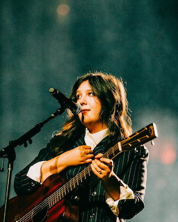Woman in pinstripe blazer and white shirt playing guitar on stage under teal lighting