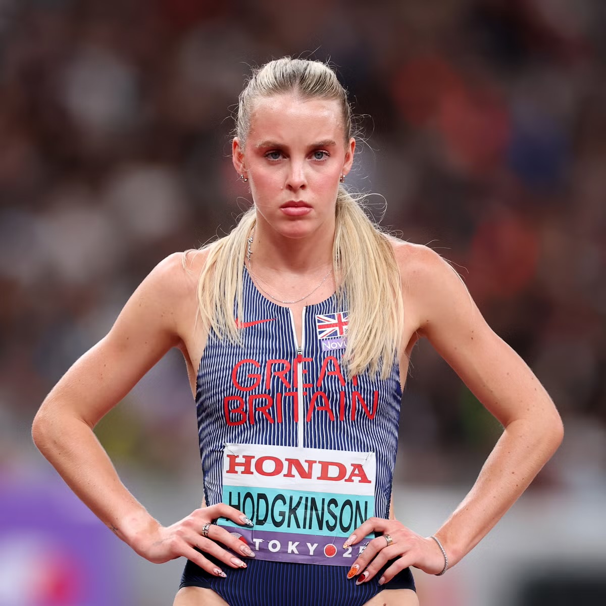 Blonde female athlete in striped competition uniform standing with hands on hips at track stadium
