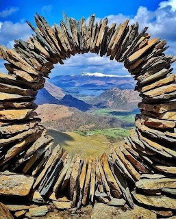 Circular dry stone arch framing a mountain valley landscape with lakes and snow-capped peaks