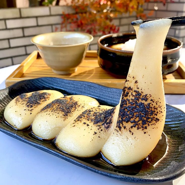 Grilled Japanese mochi rice cakes on a black plate with sauce and small bowls in the background