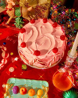Pink frosted cake with cherries on top surrounded by Christmas decorations and colorful tinsel