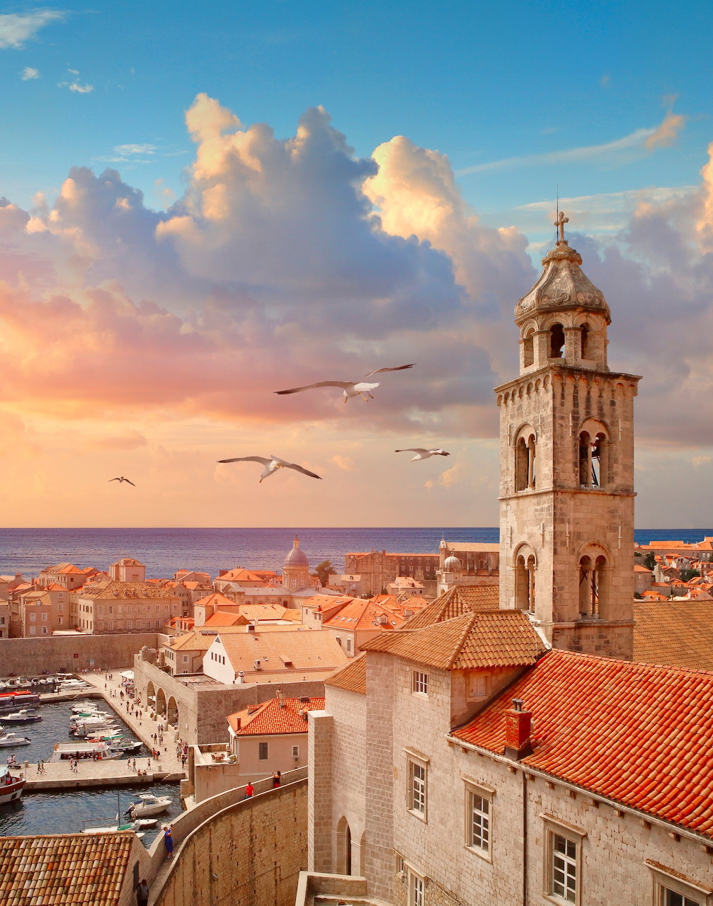 Historic coastal city at sunset with red roofs, stone bell tower, seagulls, and a marina by the sea