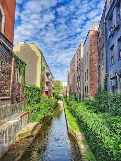 Narrow urban canal lined with ivy-covered brick buildings and green vegetation under a blue sky