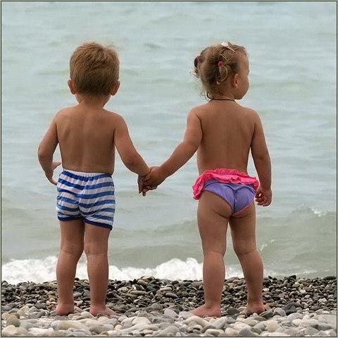 Two small children in swimsuits holding hands on a pebbly shore facing the water
