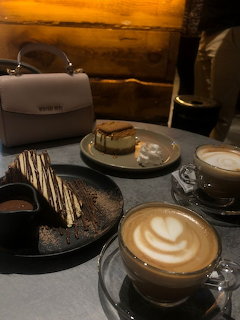 Latte with heart-shaped foam art, layered cake slice, and white purse on café table with warm lighting