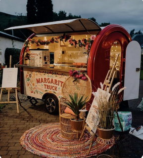 Red trailer bar with lights, signs, flowers, and potted plants on an outdoor brick patio