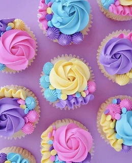 Colorful cupcakes with rose-swirl frosting and flower decorations arranged on purple surface