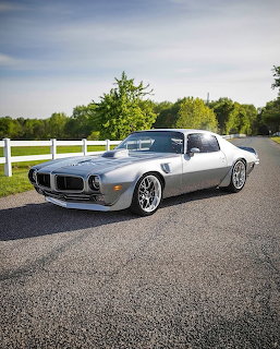 Silver classic muscle car parked on a road beside a white fence and trees