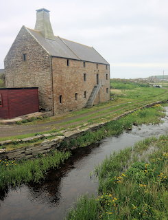 Stone watermill building with red door beside rural stream under overcast sky