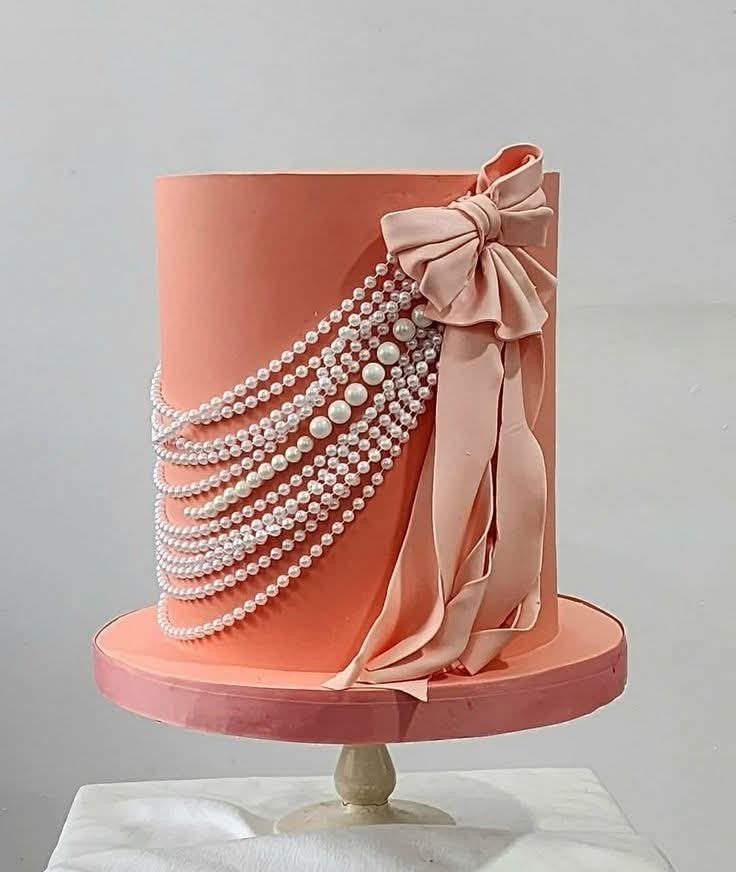 Elegant coral fondant cake with draped pearl strands and a large pink bow on a white cake stand