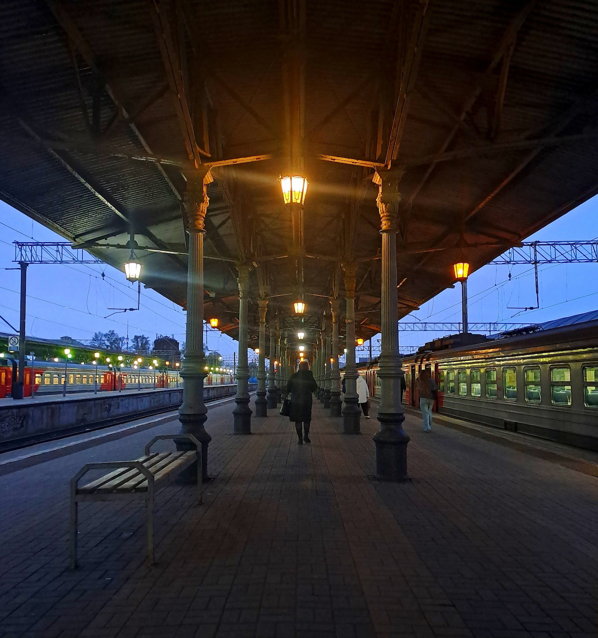 Train station platform at dusk with glowing lights, people walking, and trains on tracks under evening sky
