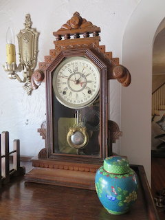 Antique wooden Waterbury calendar clock with pendulum, beside a cloisonné jar and brass wall sconce