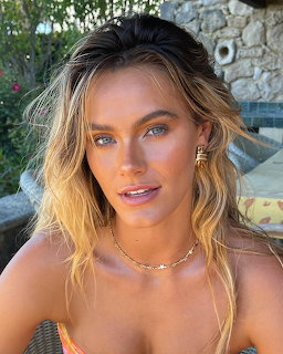 Close-up of a blonde woman with blue eyes wearing a gold necklace outdoors in sunlight