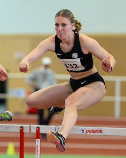 Female athlete mid-jump clearing a hurdle during an indoor track and field event