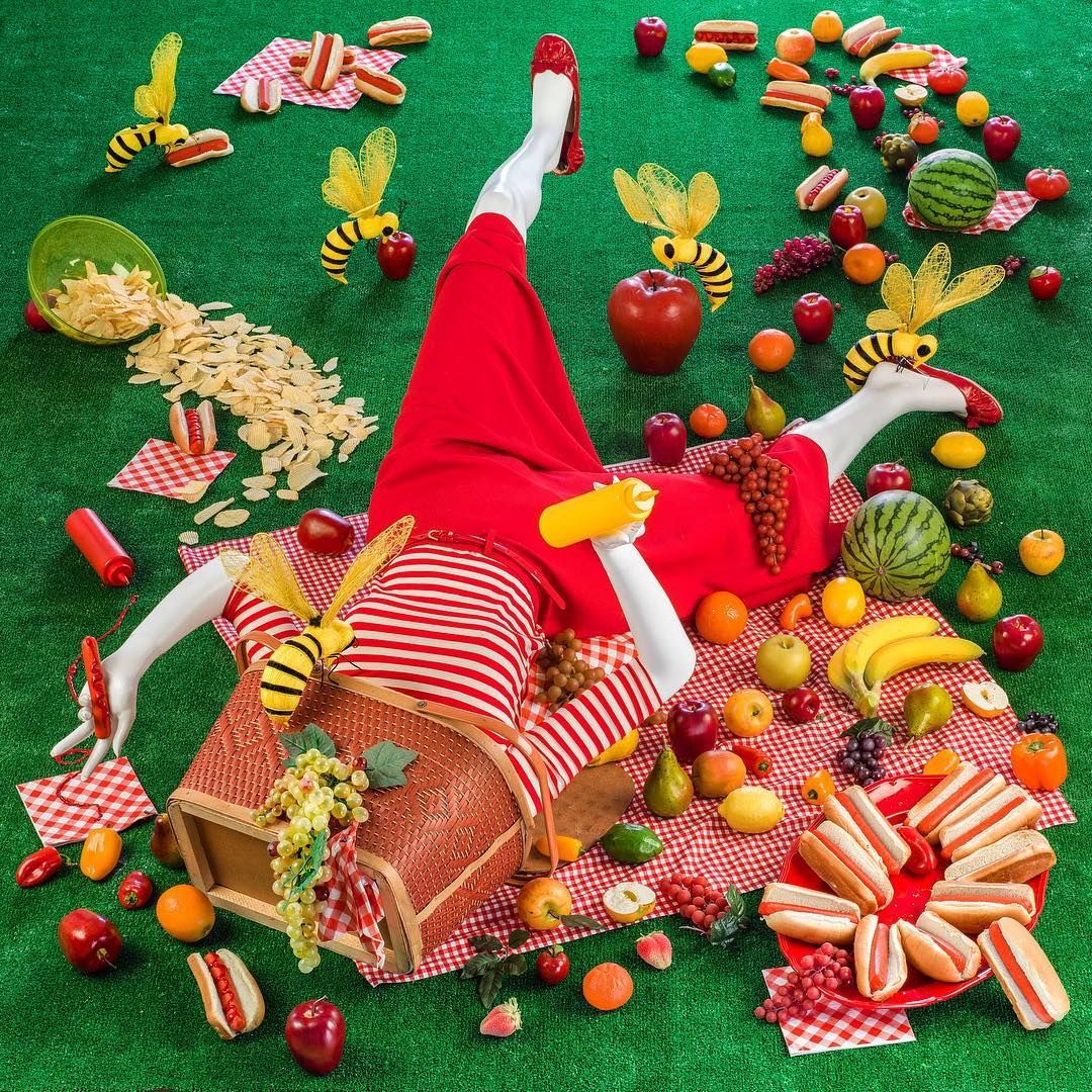 Stylized woman in a red dress lying among fruit, pastries, flowers, and bee decorations on green background