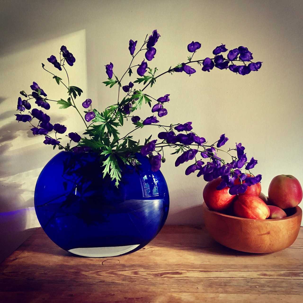 Purple flowers in a blue vase next to a bowl of red apples on a wooden surface