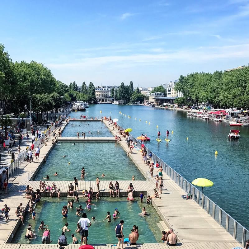 Aerial view of crowded outdoor pools beside a canal in Paris on a sunny summer day
