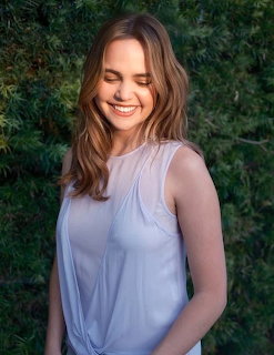Smiling young woman with wavy brown hair in a sleeveless light top standing before green foliage