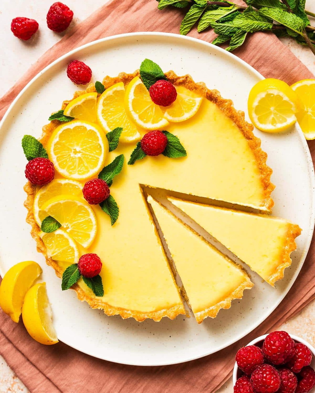 Lemon tart with slice removed, decorated with lemon slices and fresh strawberries on white plate