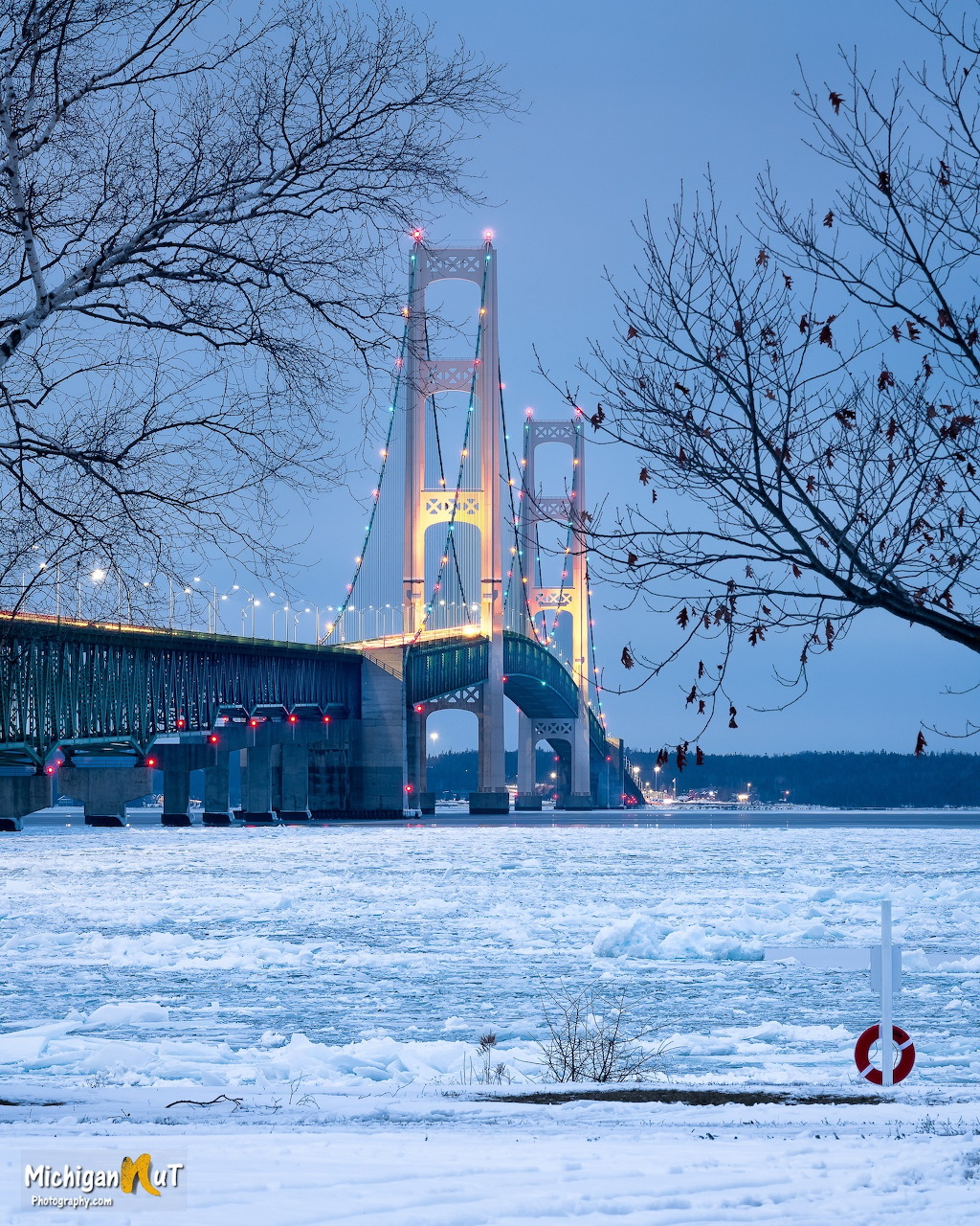 Mackinac Bridge glowing with lights over a frozen strait at dusk, surrounded by bare winter trees