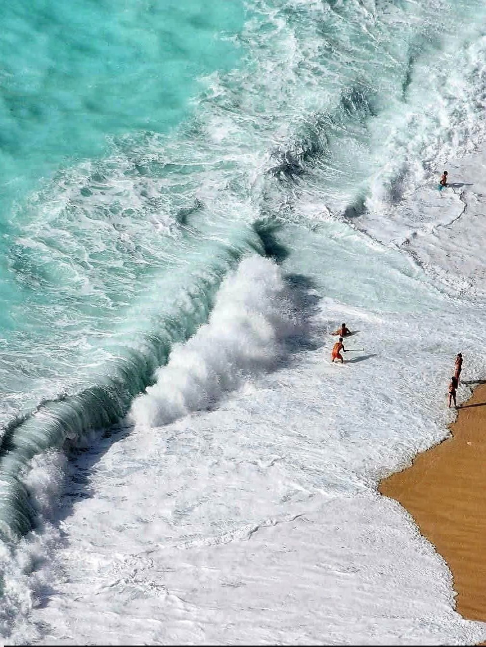 Overhead view of turquoise ocean waves breaking on white pebble beach with people