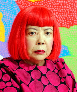 Elderly Japanese woman with bright red bob wig wearing pink and black polka dot top in front of colorful dotted art