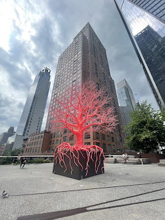 Red tree sculpture with roots on black cube in a New York City plaza with tall buildings behind
