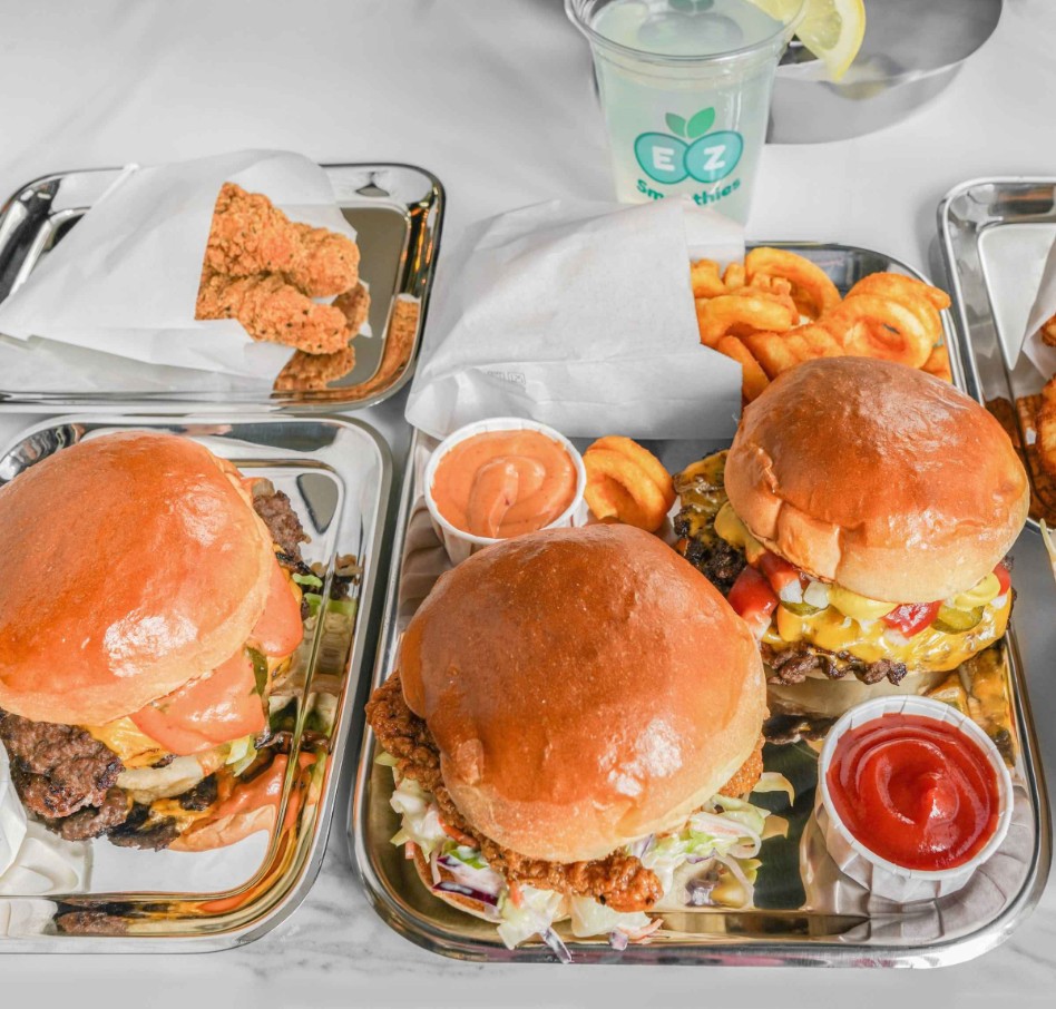 Two smashburgers with cheese and toppings on metal trays, served with fries and drinks
