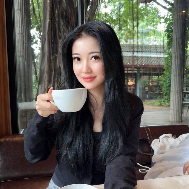 Woman with long black hair holding a white cup indoors by a window