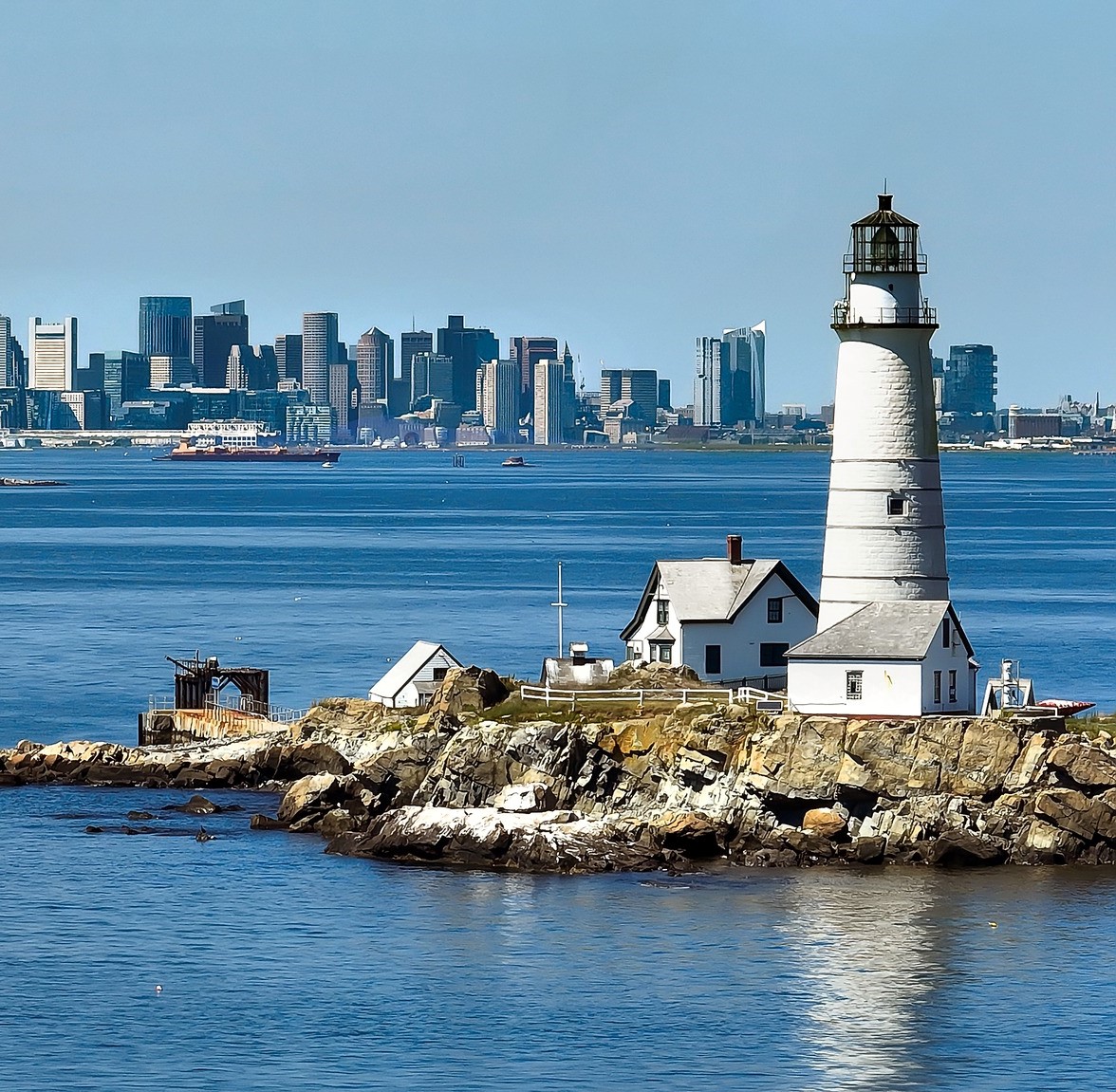 White lighthouse and house on rocky island with Boston skyline across harbor