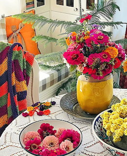 Yellow vase with pink and orange flowers on lace tablecloth beside bowls of flowers and a crocheted blanket