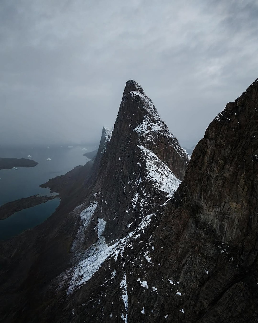 Sharp rocky mountain peak with snow patches overlooking a fjord under cloudy skies